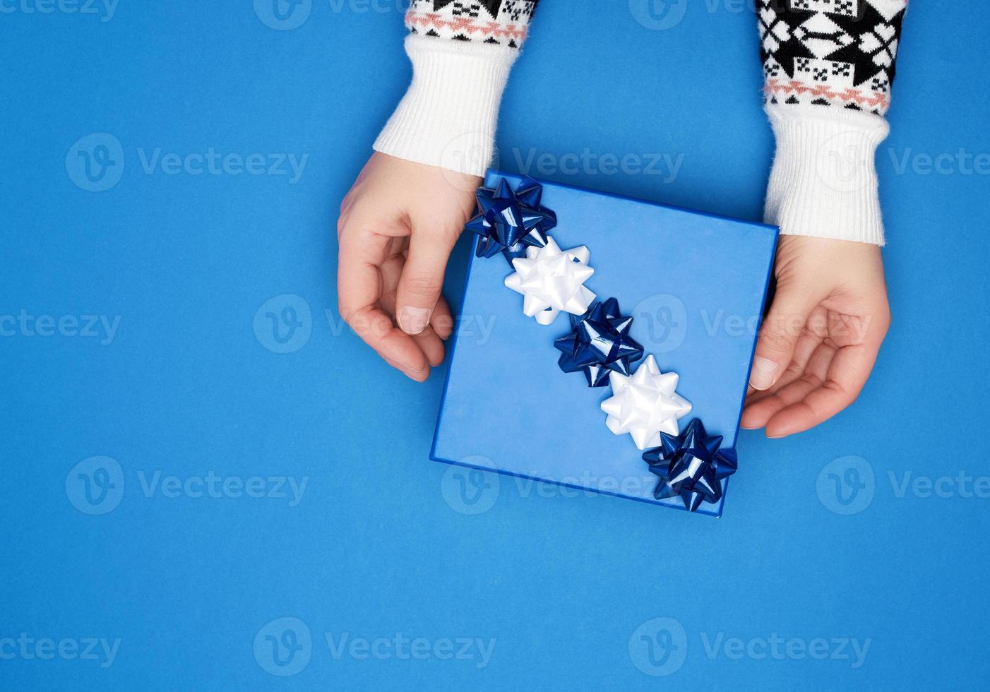 two female hands in a gray sweater hold closed square blue box with a bow photo