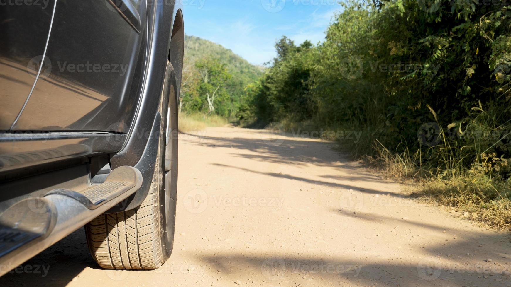 Tires of a gray car traveling and stepping on the dirt road. Dirt road with trees and grass on both sides under blue sky. photo