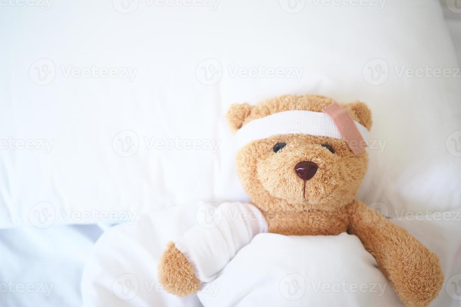 Teddy bear lying sick in bed With a headband and a cloth covered photo