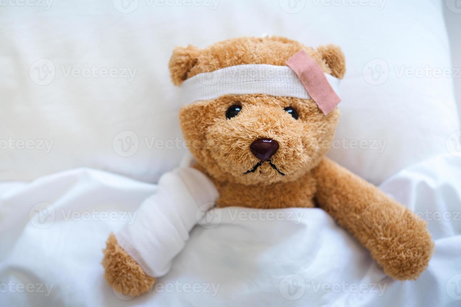 Teddy bear is sick on the bed, his arm is broken and his head is broken from an accident. photo
