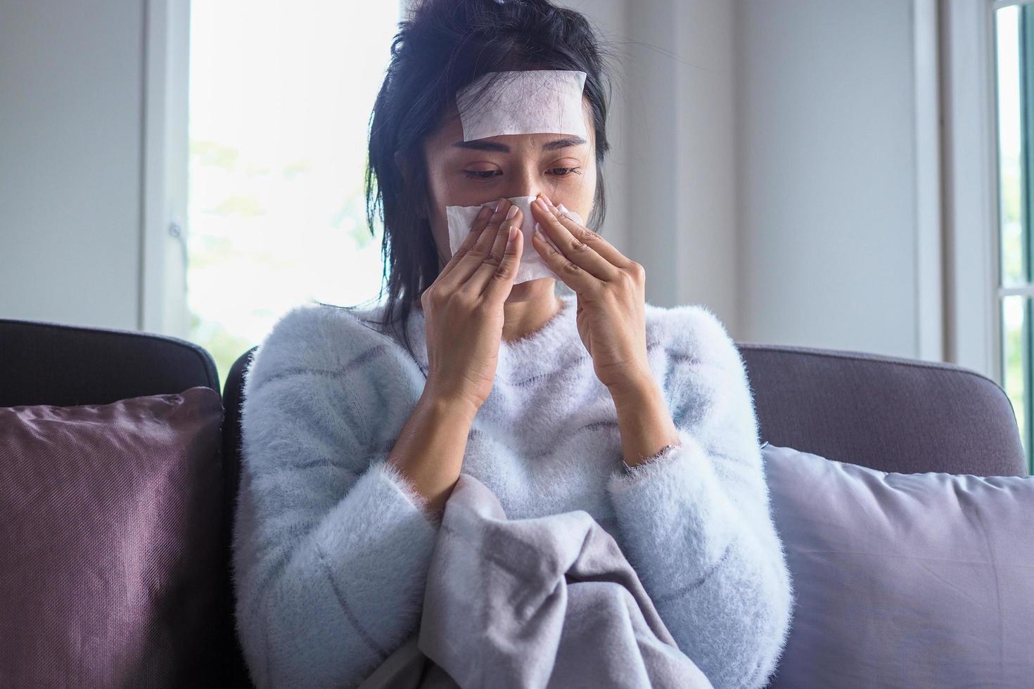 Sick woman sitting on the sofa in the house, having high fever on the forehead with a gel pad to reduce body temperature, sore throat, runny nose and coughing. photo