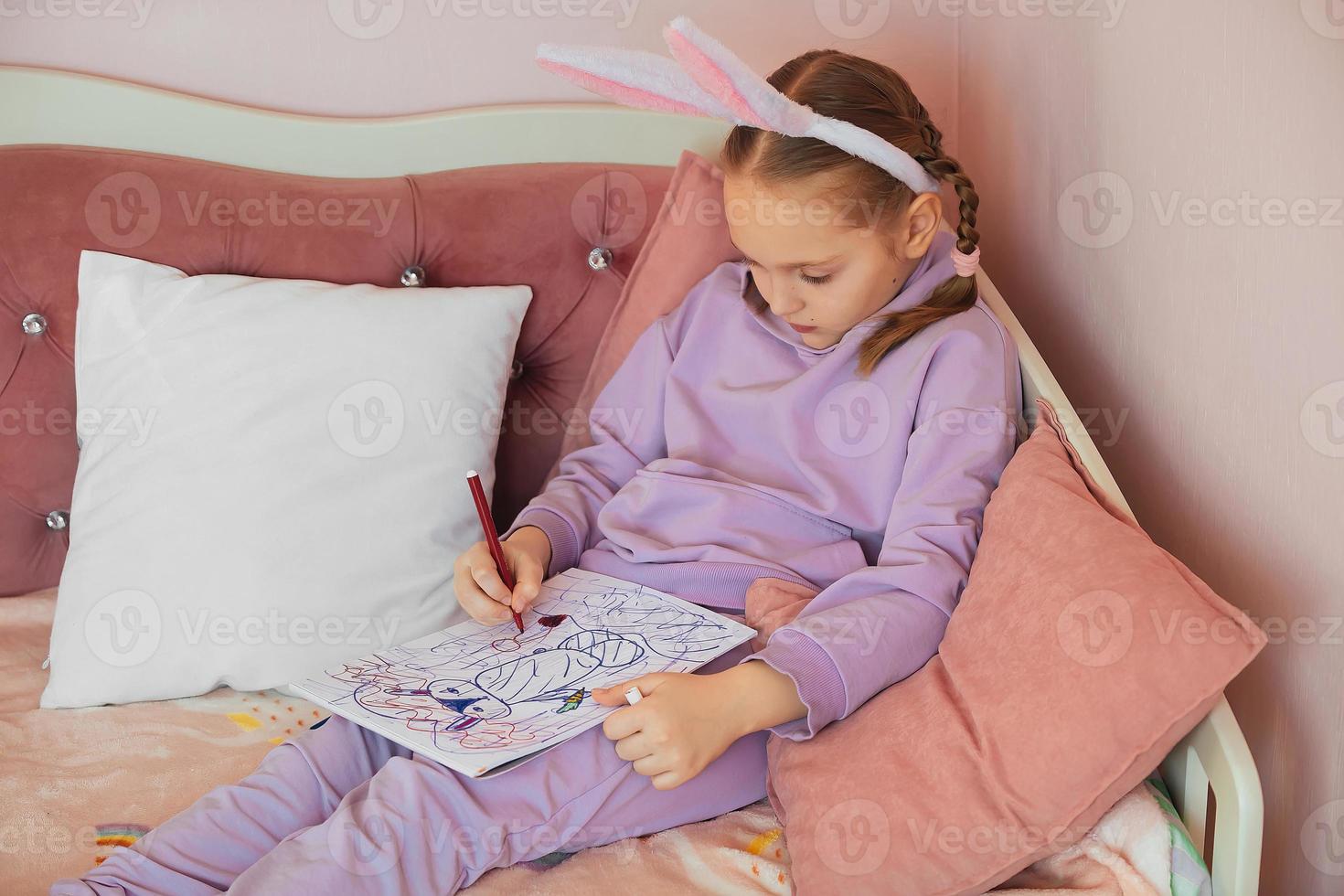 a schoolgirl girl with bunny ears on her head sits on a bed in her room and draws a bunny in an album, preparing for Easter photo