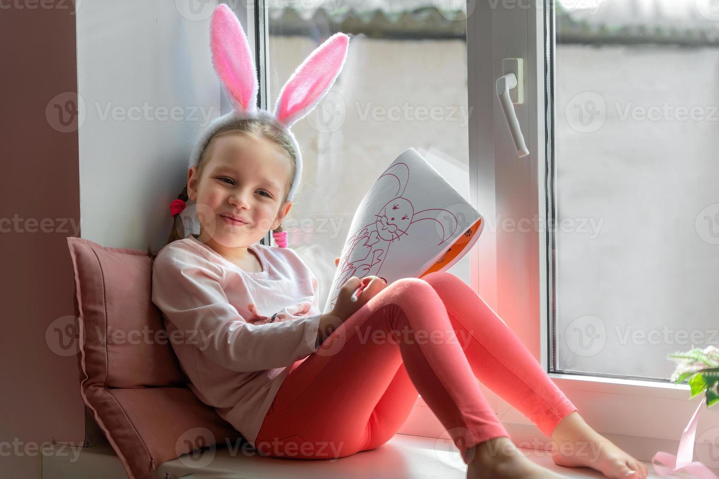 a little girl in bunny ears sits on the window and draws a bunny in her album, preparing for Easter photo