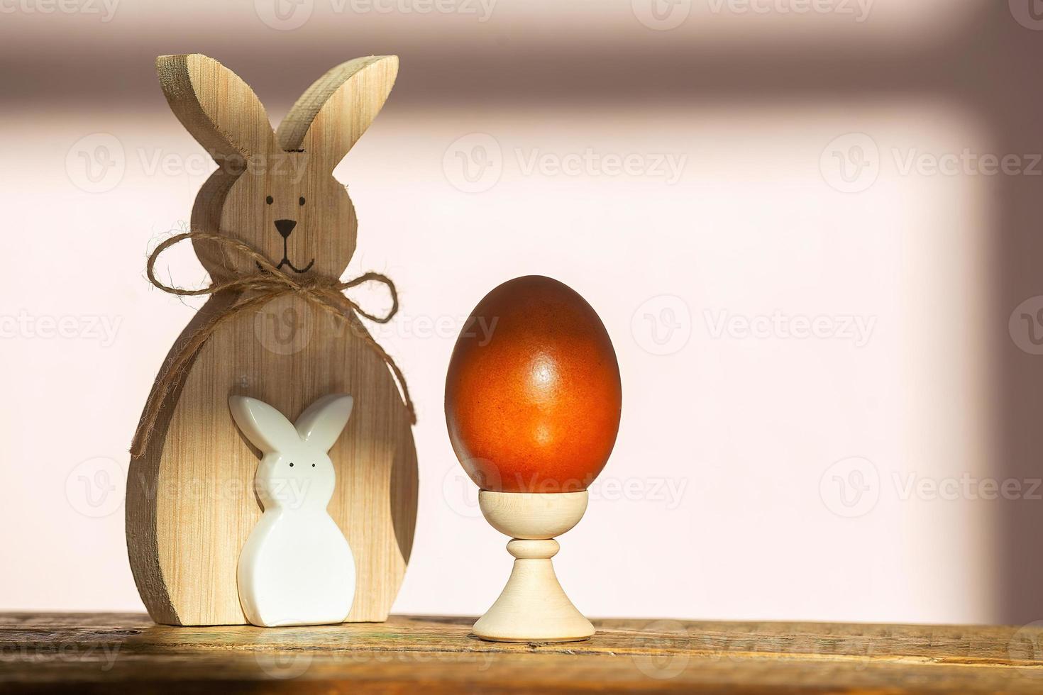 on a wooden background, there is one painted Easter red egg on a stand with natural dyes and an Easter bunny in the rays of the bright sun, minimalism, copy space. Preparing to celebrate Easter photo