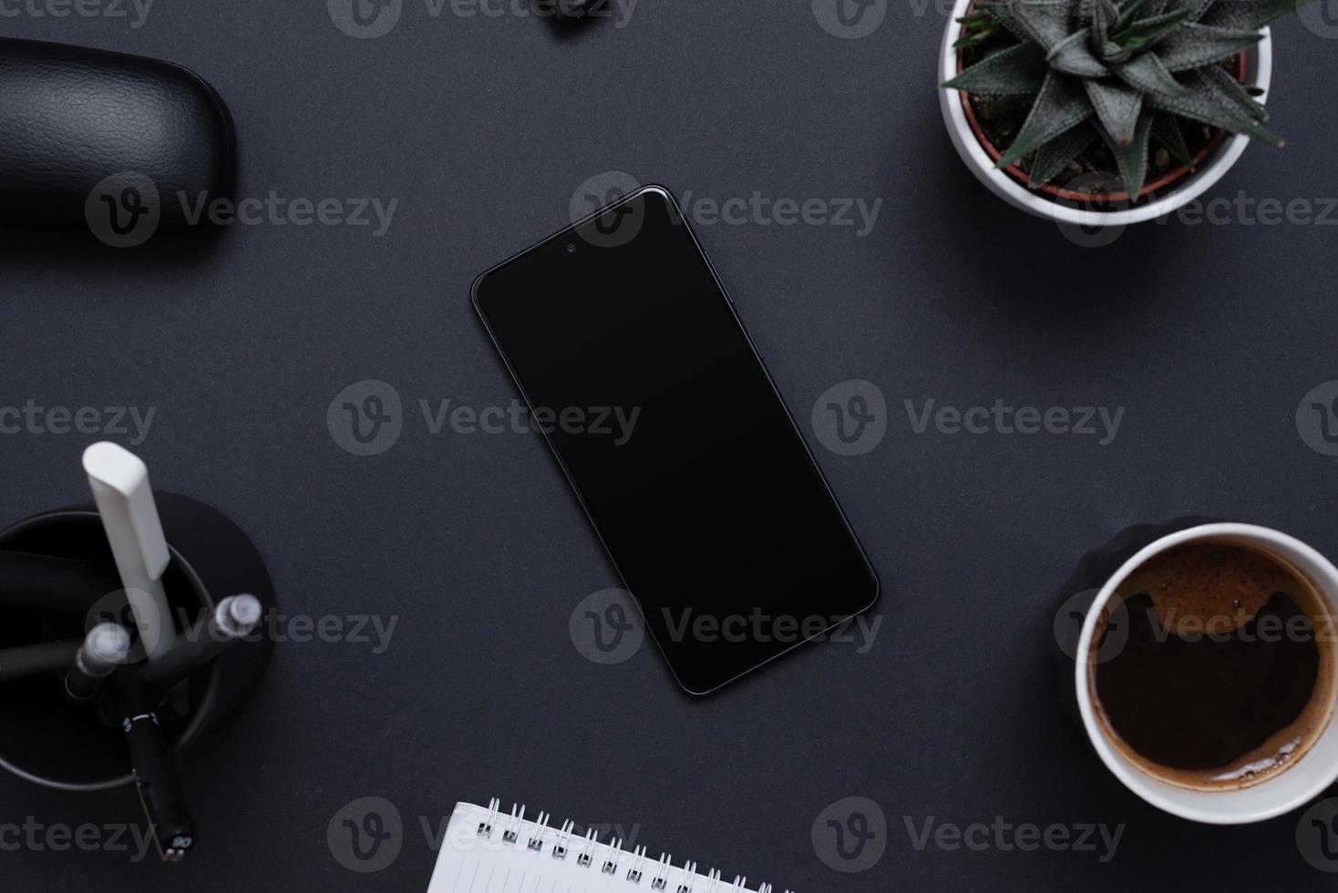 Minimalist and organized work desk with a blank smart phone surrounded by a plant, pens, coffee and notepad. Blank screen for mockup app presentation. Office work desk composition, top view, flat lay photo
