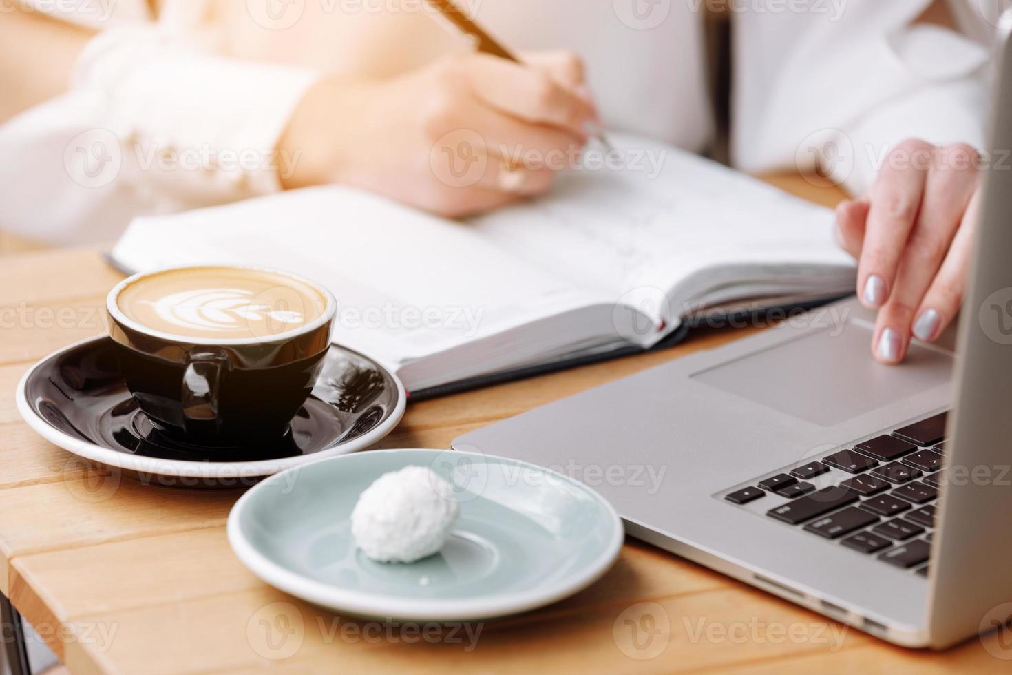 cropped photo of woman hands is doing notes in paper notebook. On table is laptop, cup of coffee and some candy. Freelancer working in coffee shop. Student learning online. Business concept