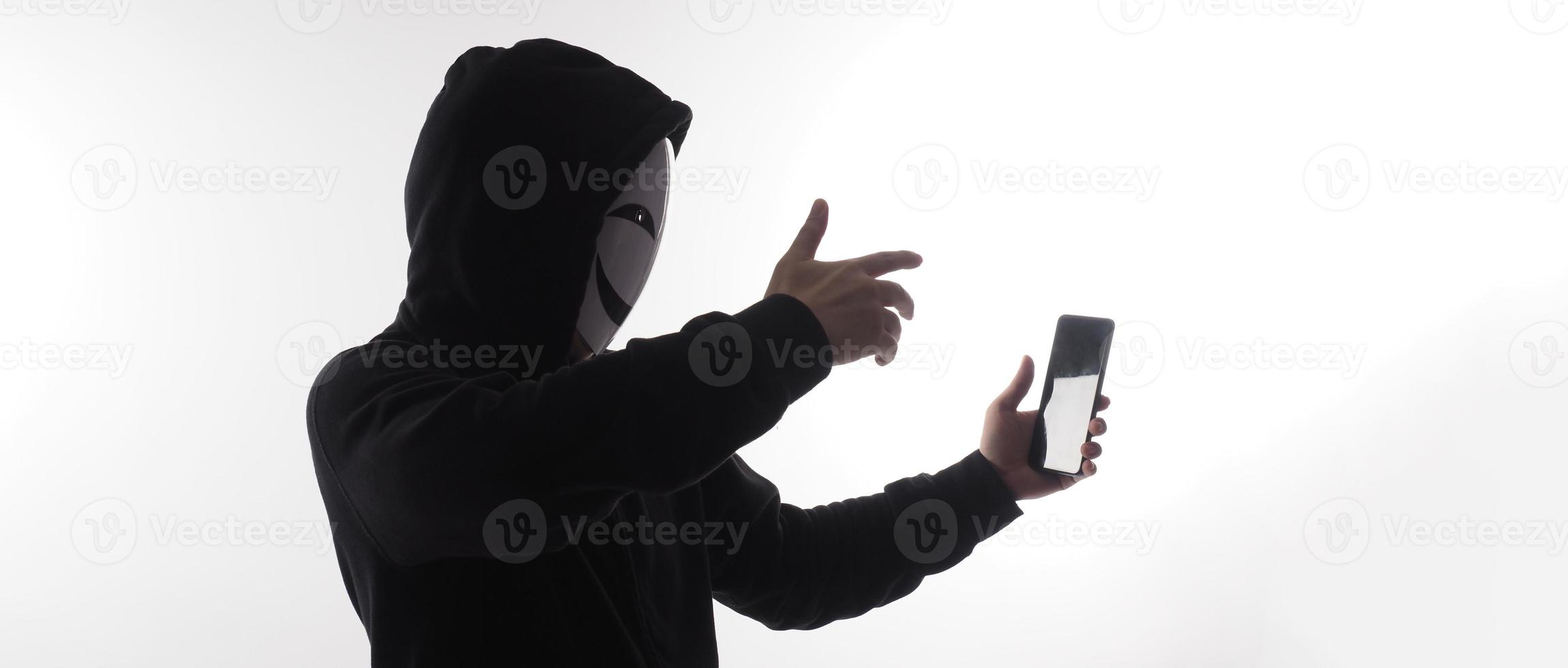 Hacker Anonymous and face mask with smartphone in hand. Man in black hood shirt holding and using mobile phone on white background. Represent cyber crime data hacking or stealing personal data concept photo