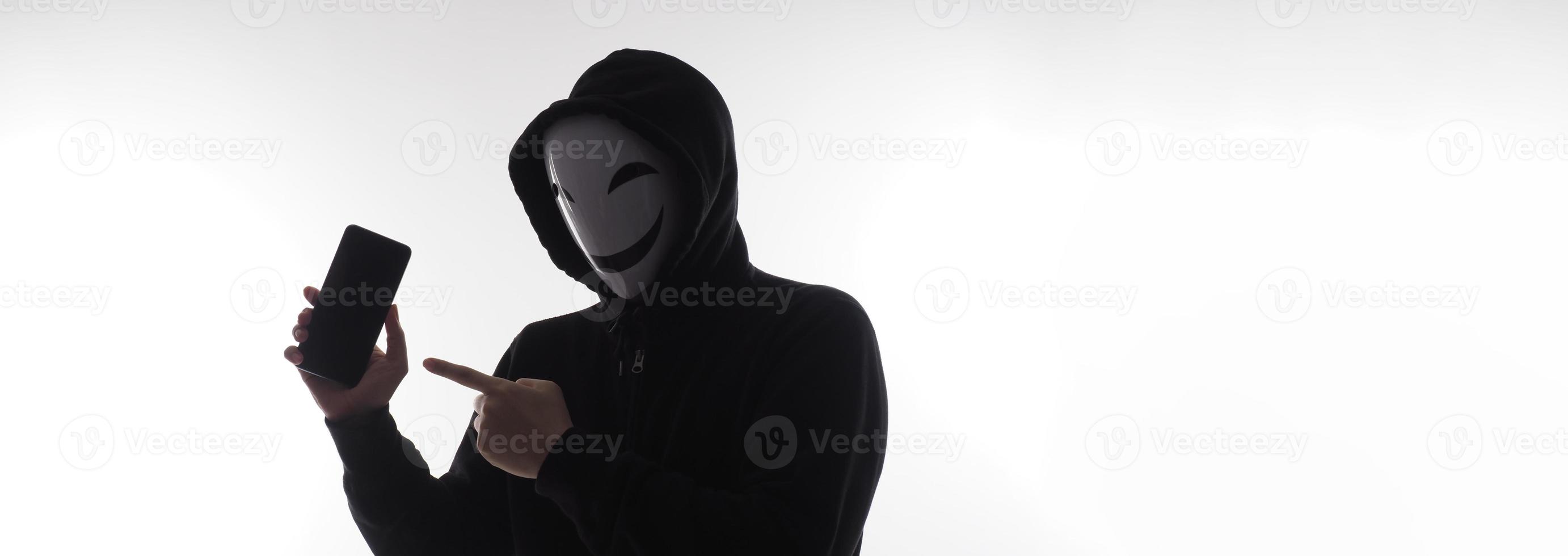 Hacker Anonymous and face mask with smartphone in hand. Man in black hood shirt holding and using mobile phone on white background. Represent cyber crime data hacking or stealing personal data concept photo