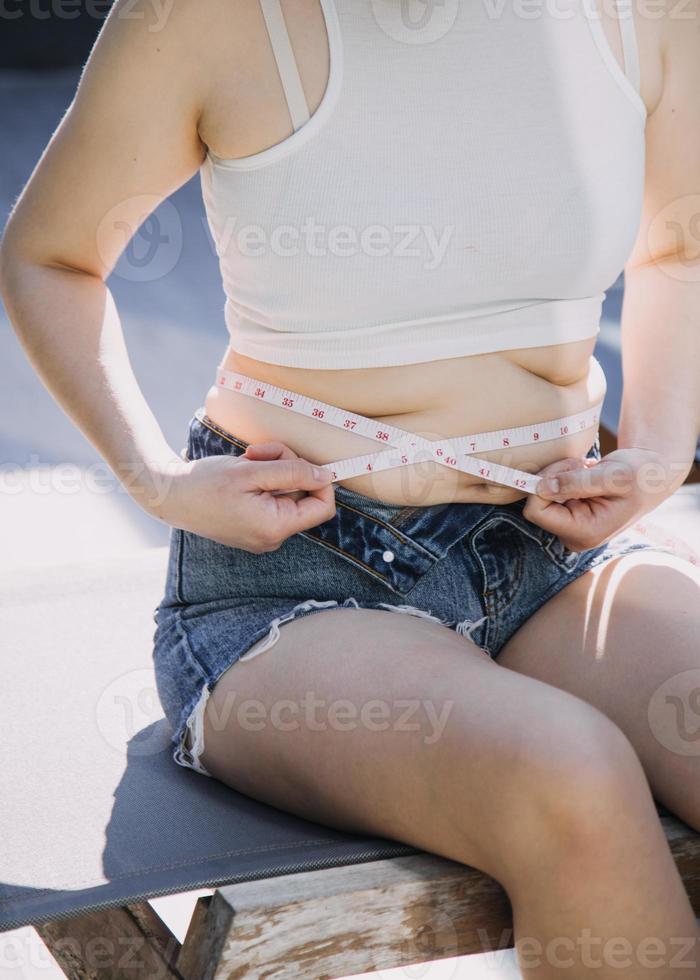 mujer gorda, barriga gorda, gordita, mujer obesa mano sosteniendo grasa abdominal excesiva con cinta métrica, concepto de estilo de vida de dieta de mujer foto