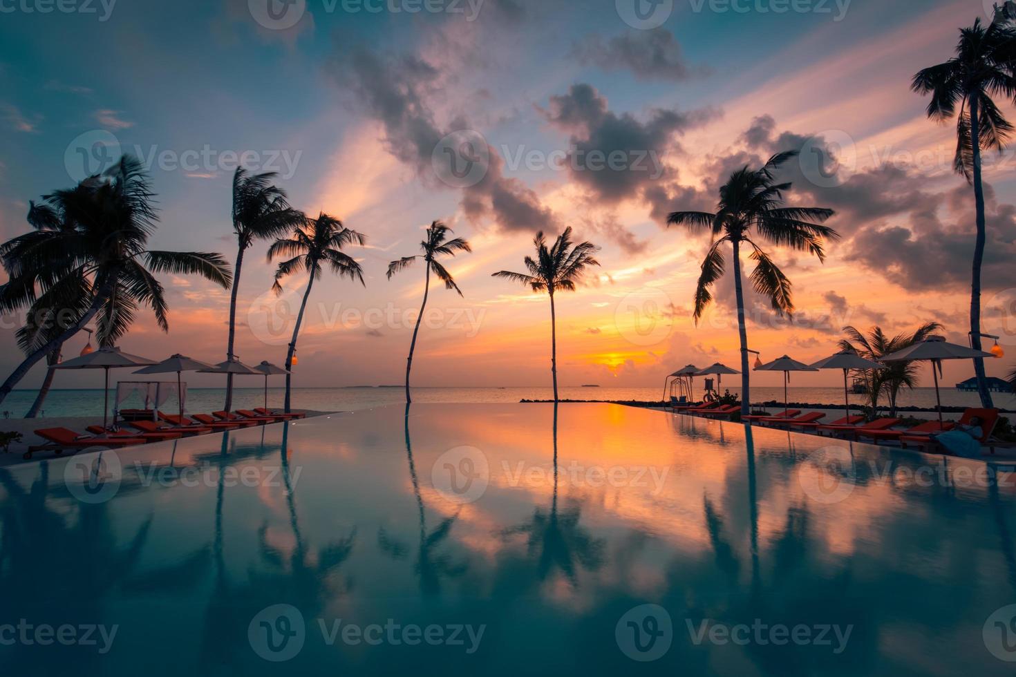 Beautiful poolside and sunset sky. Luxurious tropical beach landscape, deck chairs and loungers and water reflection. Palm trees reflection, amazing luxury summer beach landscape. Beach sunset photo