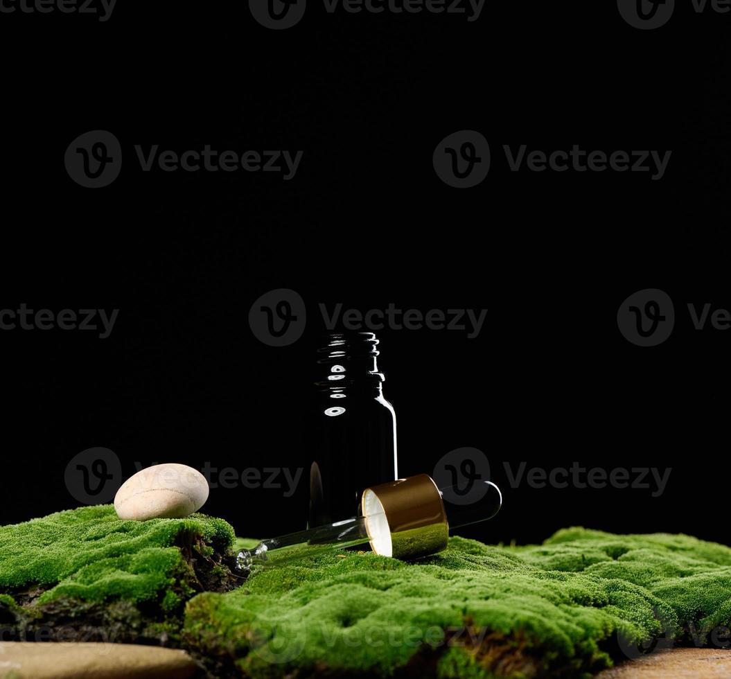 botella de vidrio negro con soportes de pipeta sobre musgo verde, fondo negro. marca de spa de cosméticos. embalaje para gel, suero, publicidad y promoción de productos, maqueta foto