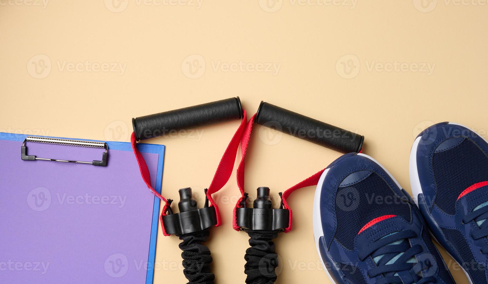 pair of blue textile sneakers, black expander and blank sheet of paper on a beige background, top view photo