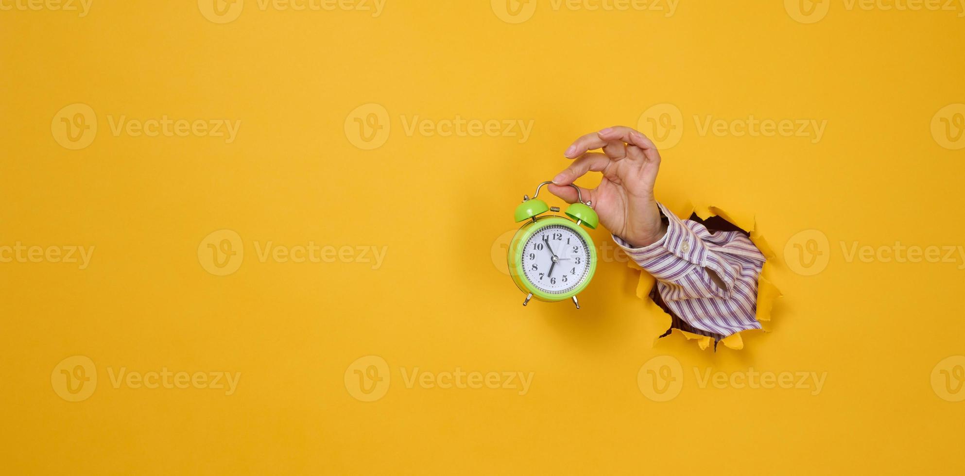 woman's hand holds a round green alarm clock, the time is five minutes to seven in the morning. A part of the body is sticking out of a torn hole in a yellow paper background. photo