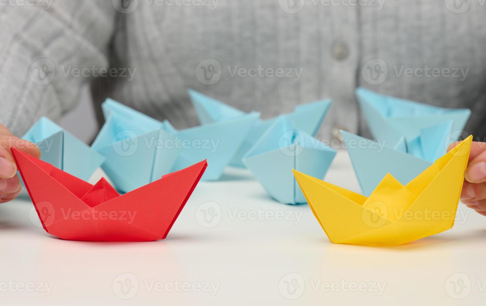female hands hold red and yellow paper boats on a white table. Confrontation, follow a strong leader, dailog photo
