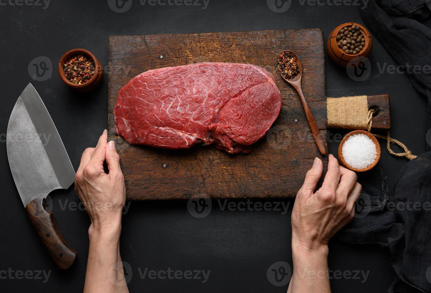 Raw beef tenderloin lies on a wooden cutting board and spices for cooking on a black table, top view and large kitchen knife photo