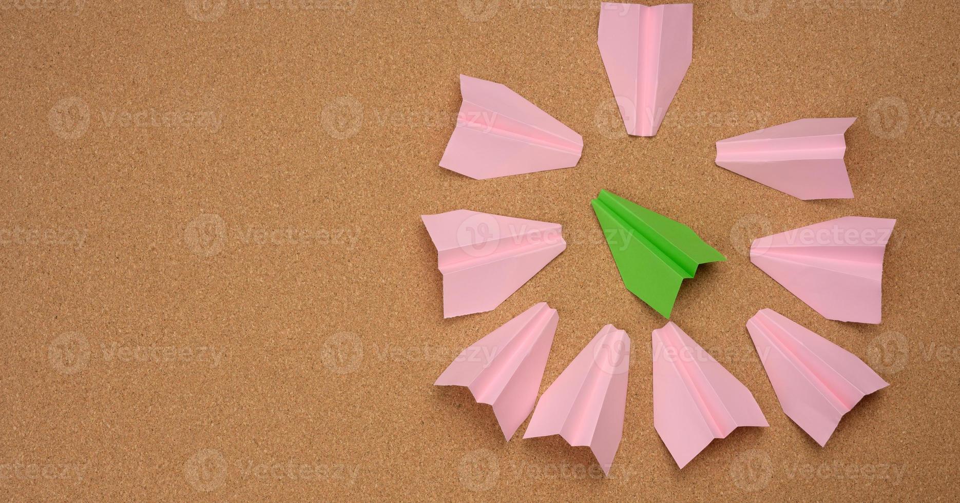green paper airplane surrounded in a ring by pink paper airplanes on a brown background photo