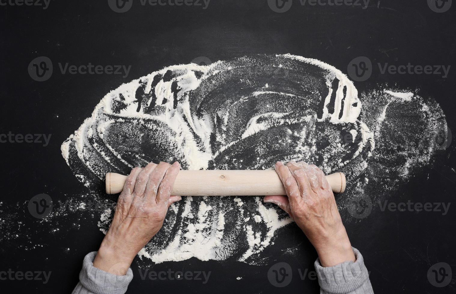 Sifted white wheat flour on a black table and two female hands hold a wooden rolling pin, top view. cooking at home photo