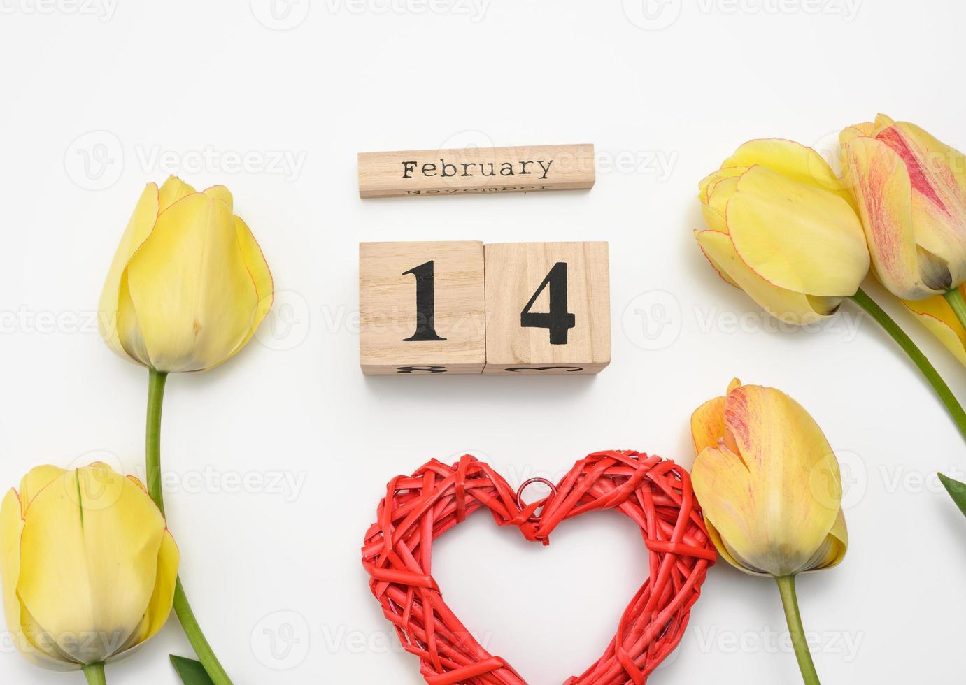 tulipanes amarillos, calendario de madera con fecha 14 de febrero y corazón rojo sobre fondo blanco foto