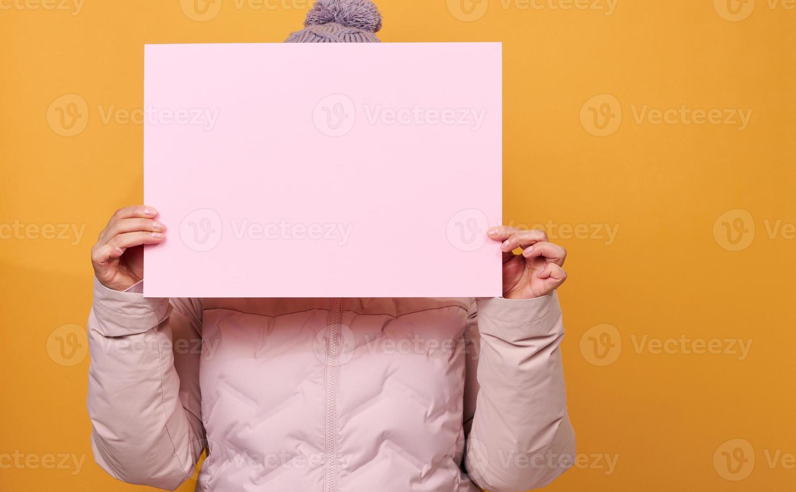 woman in pink winter jacket and hat holds blank pink sheet of paper on yellow background. Seasonal sale photo