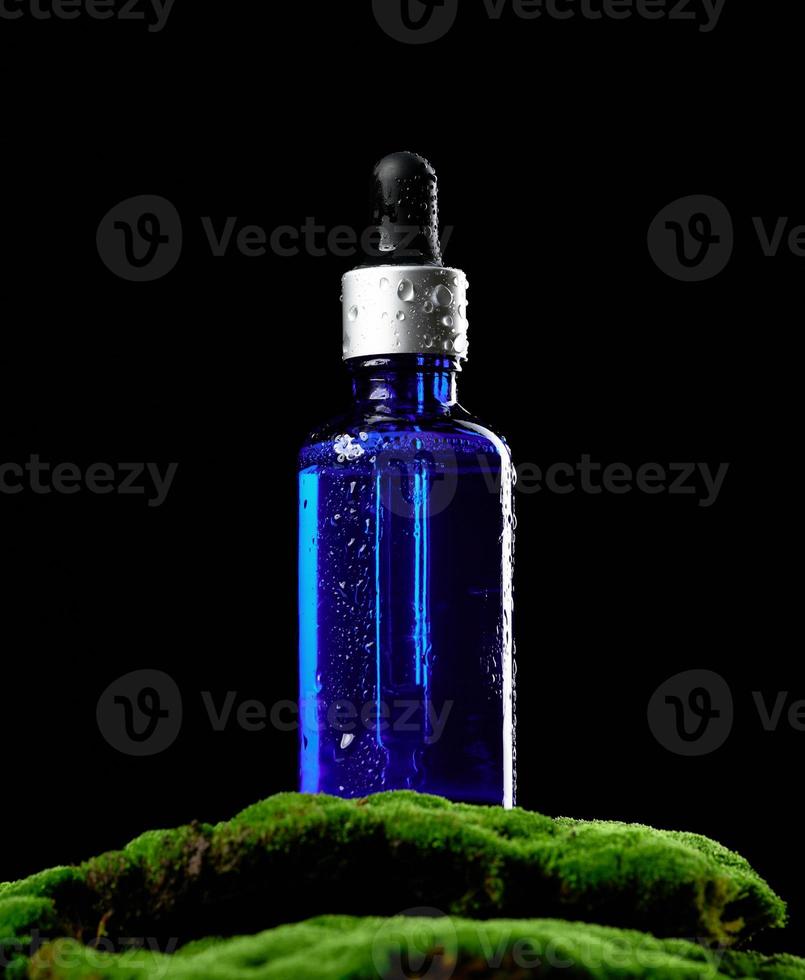 botella de vidrio azul con soportes de pipeta sobre musgo verde, fondo negro. maqueta de marca de spa de cosméticos foto