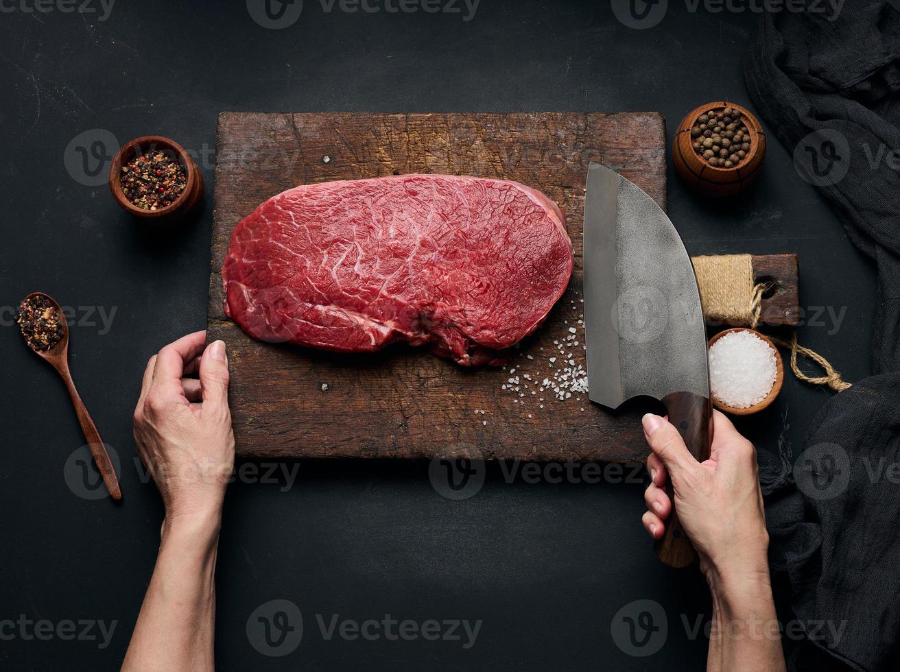 Raw beef tenderloin lies on a wooden cutting board and spices for cooking on a black table, top view photo