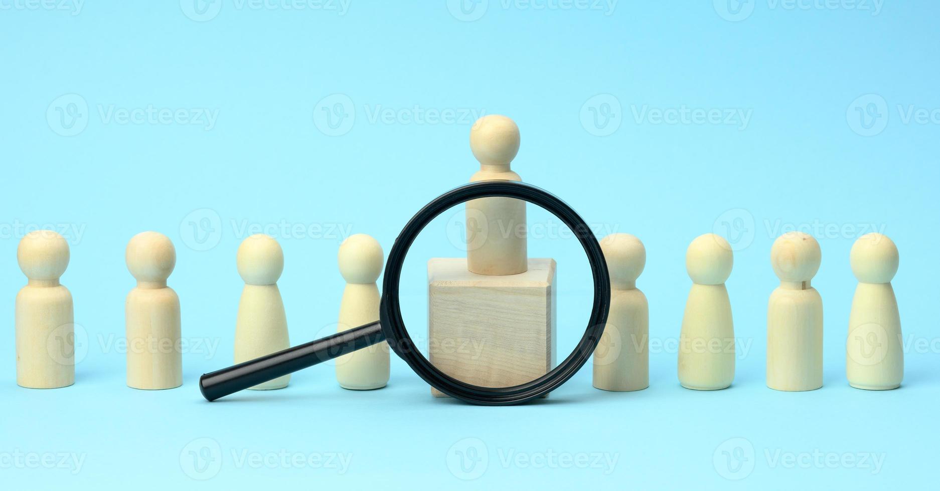 wooden figures of men stand on a blue background and a black magnifying glass. Recruitment concept, photo