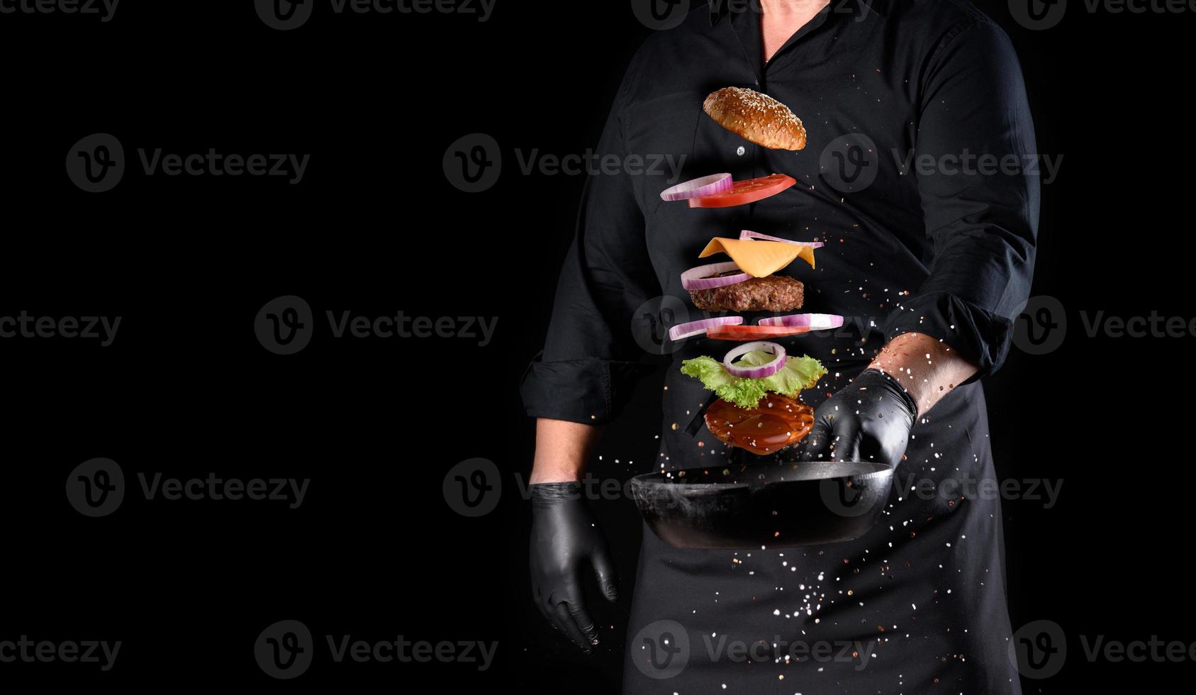 hombre con uniforme negro sosteniendo una sartén redonda de hierro fundido con ingredientes de hamburguesa con queso levitando foto