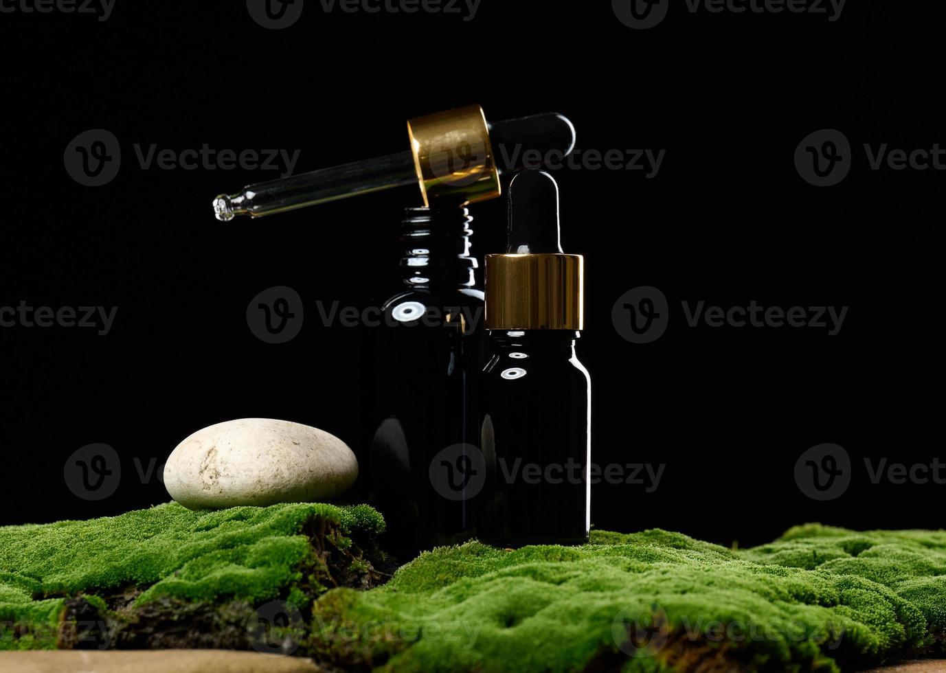 black glass bottle with pipette stands on green moss, black background. Cosmetics SPA branding. Packaging for gel, serum, advertising and product promotion, mock up photo