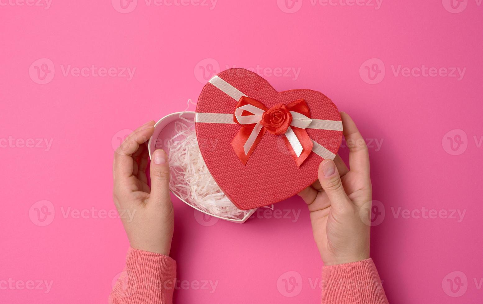 two female hands hold a red heart-shaped box on a pink background. Festive backdrop photo