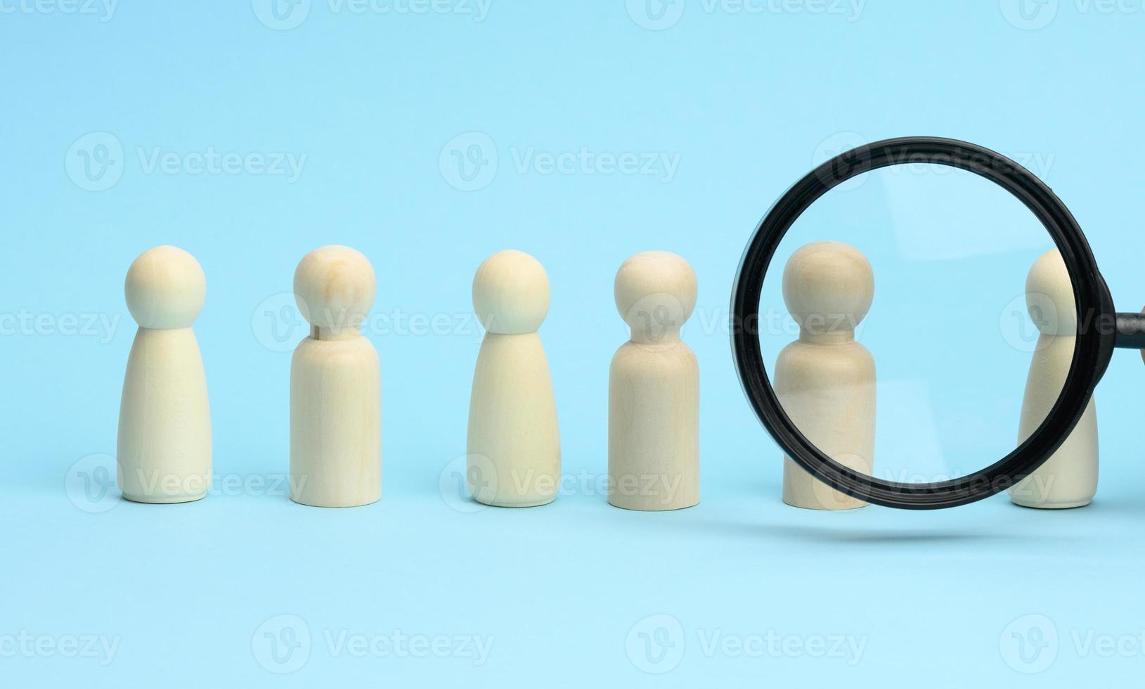 wooden figures of men stand on a blue background and a black magnifying glass. Recruitment concept, search for talented and capable employees photo