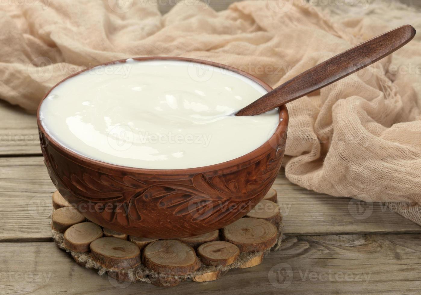 sour cream in a brown ceramic bowl with a wooden spoon on a wooden background, fermented milk useful product photo