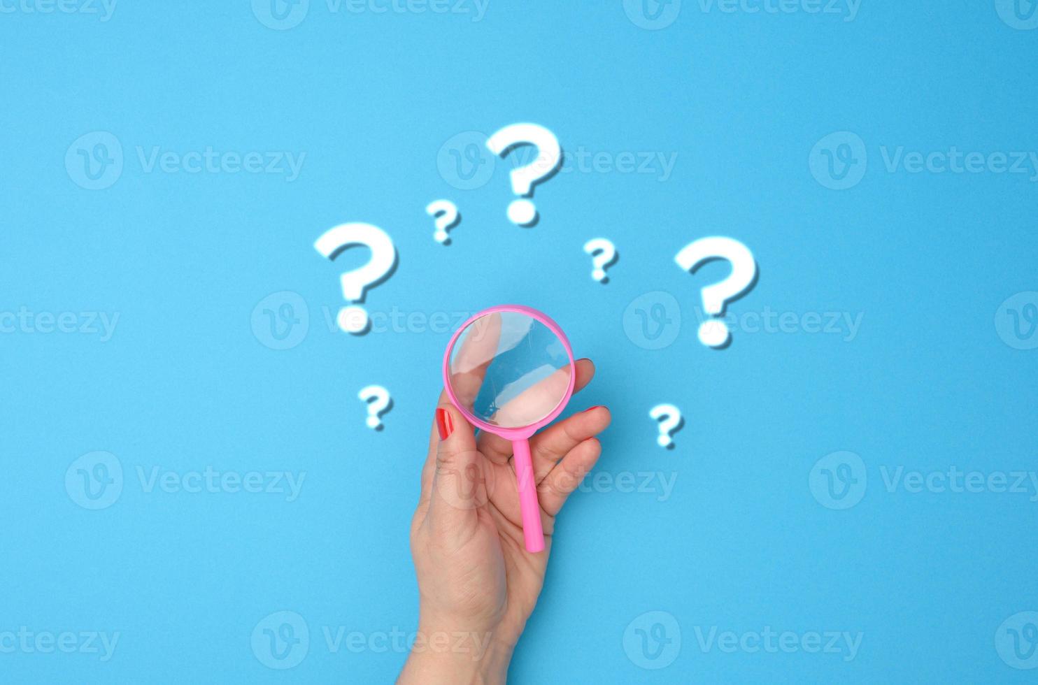 female hand holds a plastic magnifying glass and question marks on a blue background photo