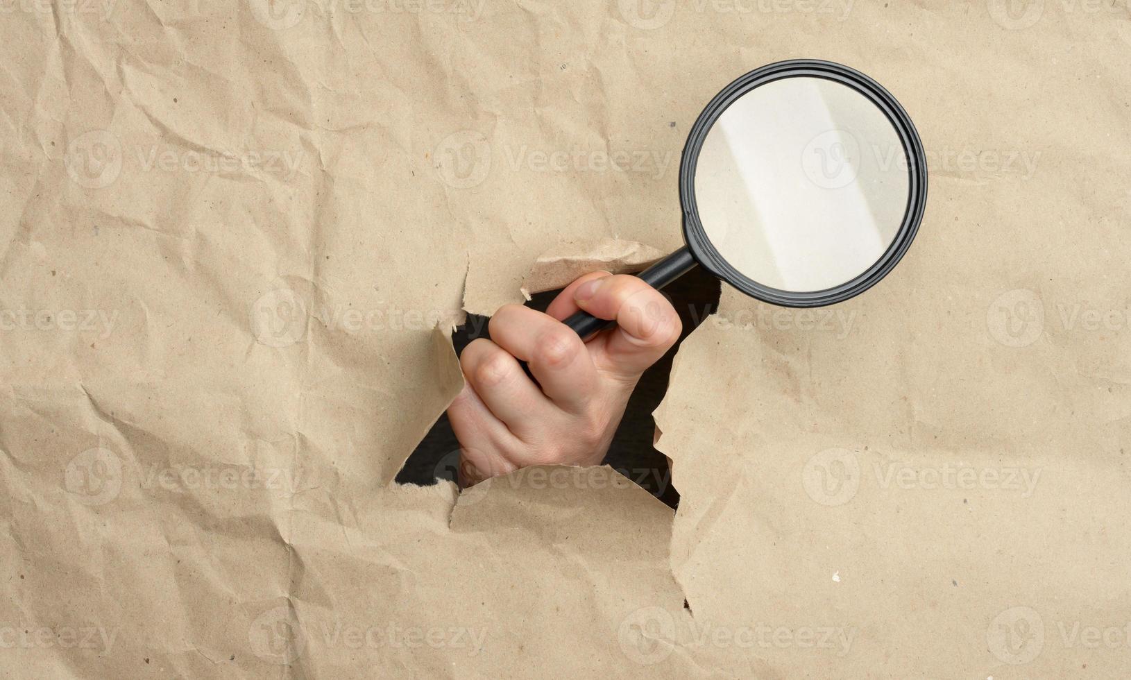 a woman's hand holds a glass magnifier, part of the body sticks out of a hole in brown paper photo