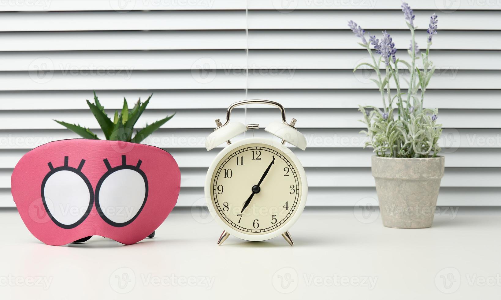 pink textile mask and round white alarm clock on the table, behind the white blinds photo