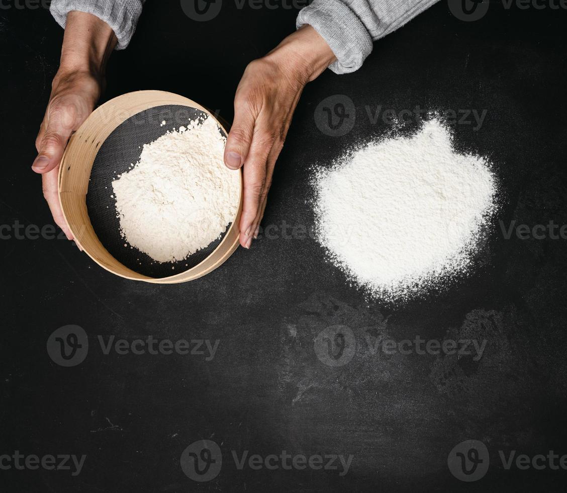 two female hands hold a round wooden sieve with wheat flour on a black table, top view photo