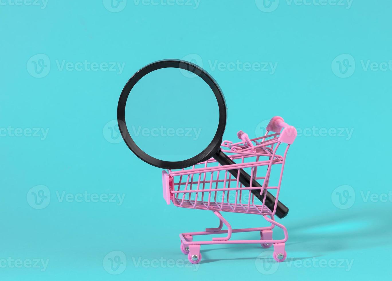carro rosa de metal en miniatura y lupa de plástico negro sobre un fondo azul claro. el concepto de búsqueda y selección de compras foto