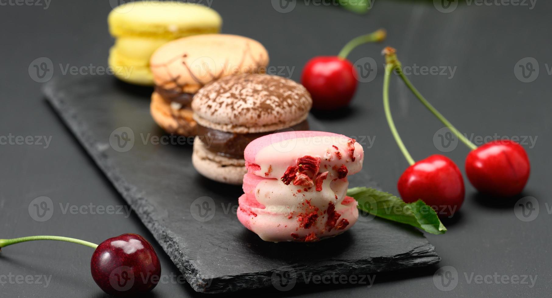 multicolored macarons and ripe red cherries on black wooden background photo