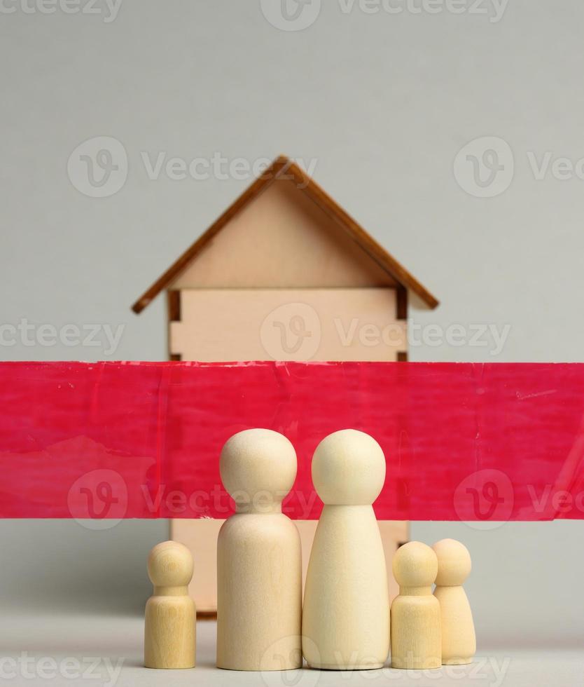 family of wooden figures and a wooden house behind a red ribbon on a gray background. Quarantine concept photo