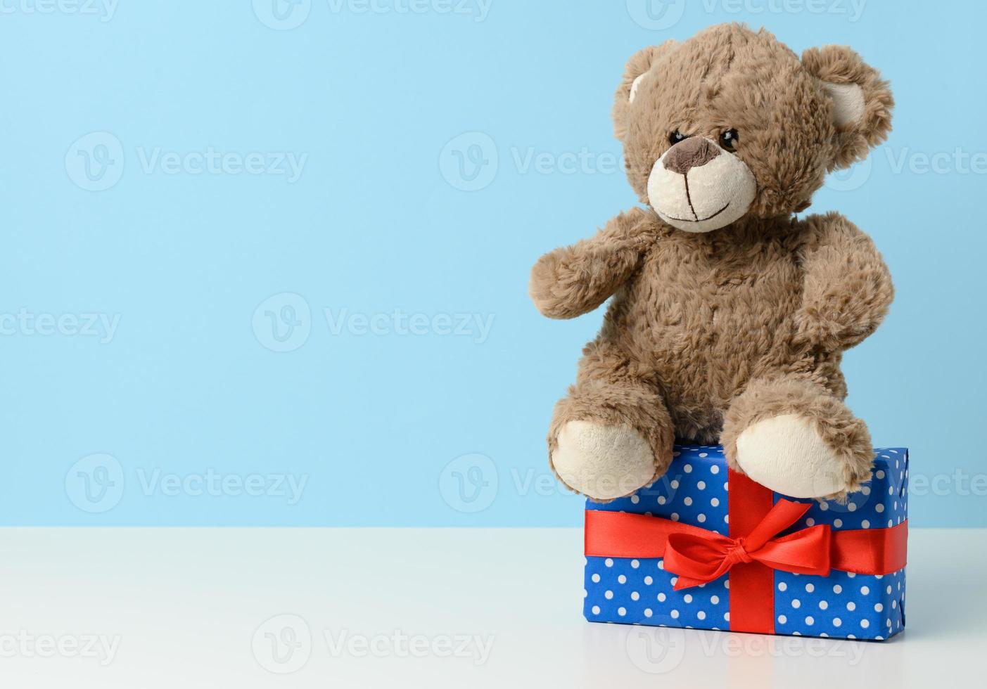 cute brown teddy bear holding a box wrapped in blue paper and red silk ribbon on white table. Prize and congratulations photo