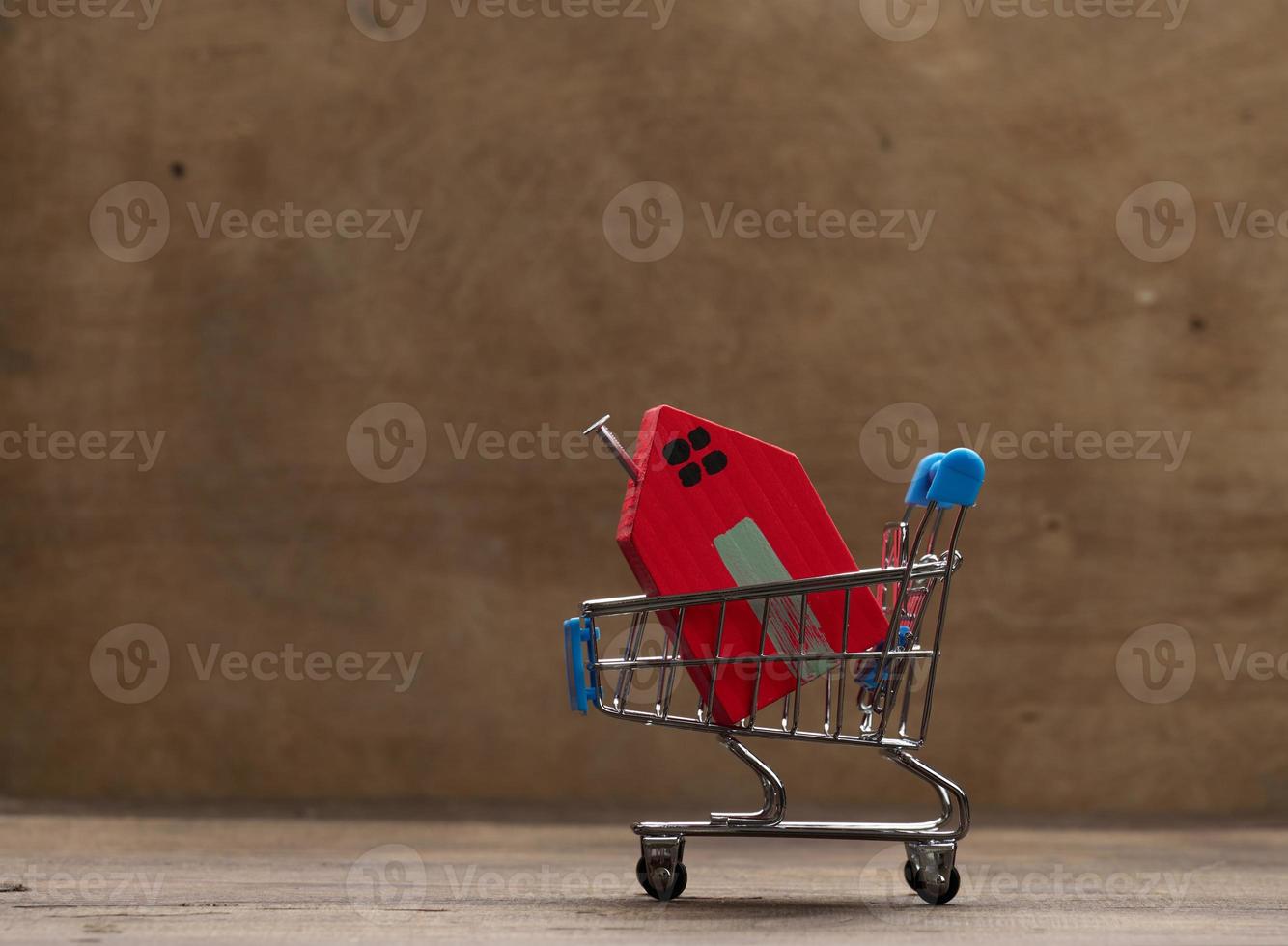 red wooden house in a metal miniature cart on a brown background. Real estate purchase concept photo