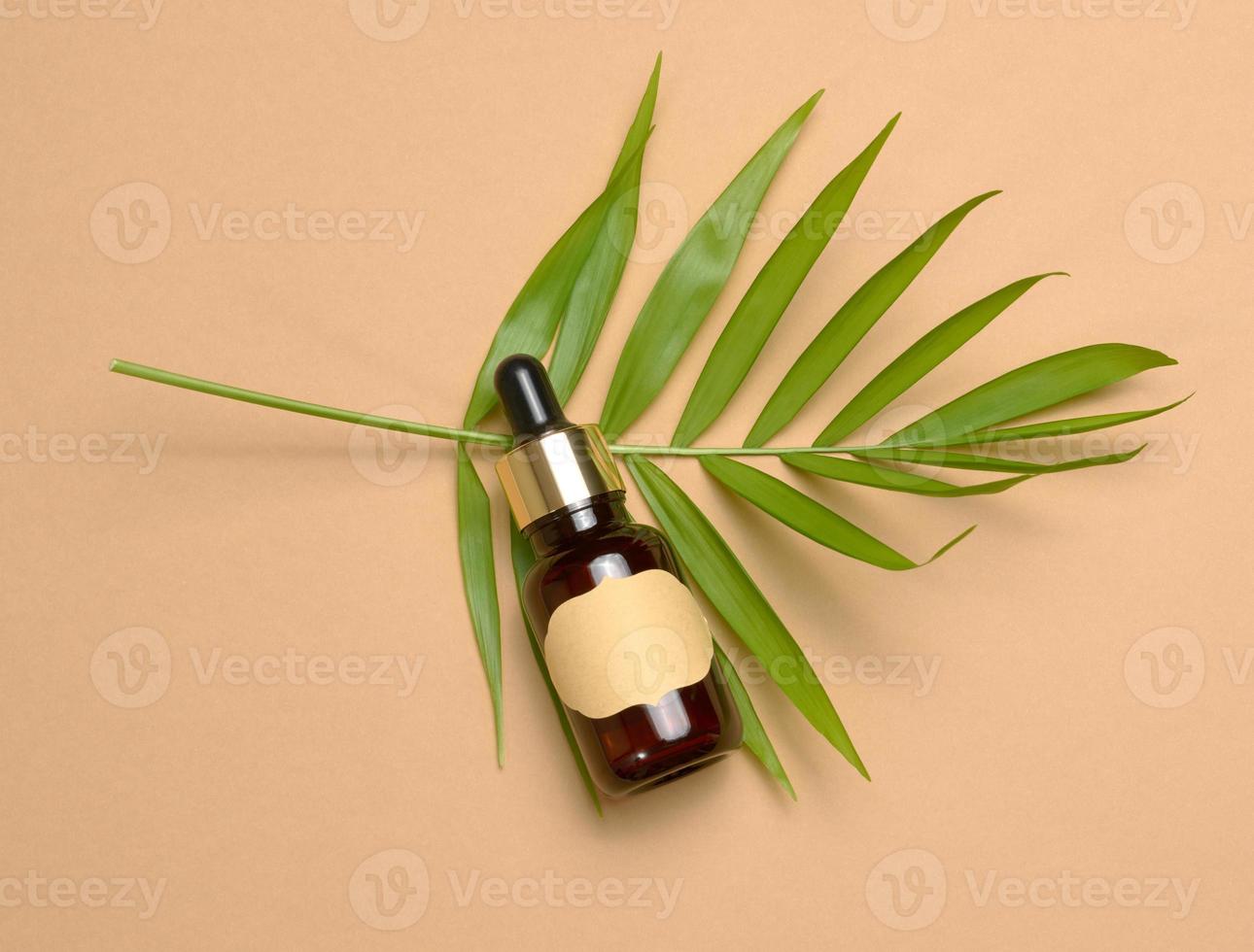 botella de vidrio marrón con una pipeta y con una etiqueta de papel en blanco sobre un fondo marrón. recipiente para cosméticos, suero y aceite, hoja de palma foto