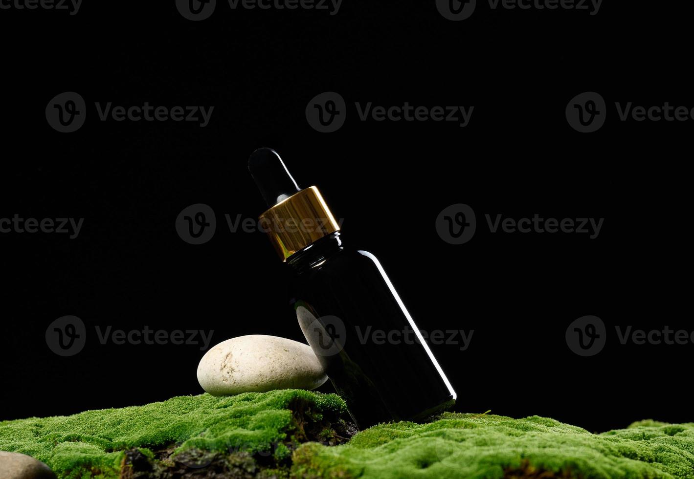 black glass bottle with pipette stands on green moss, black background. Cosmetics SPA branding photo