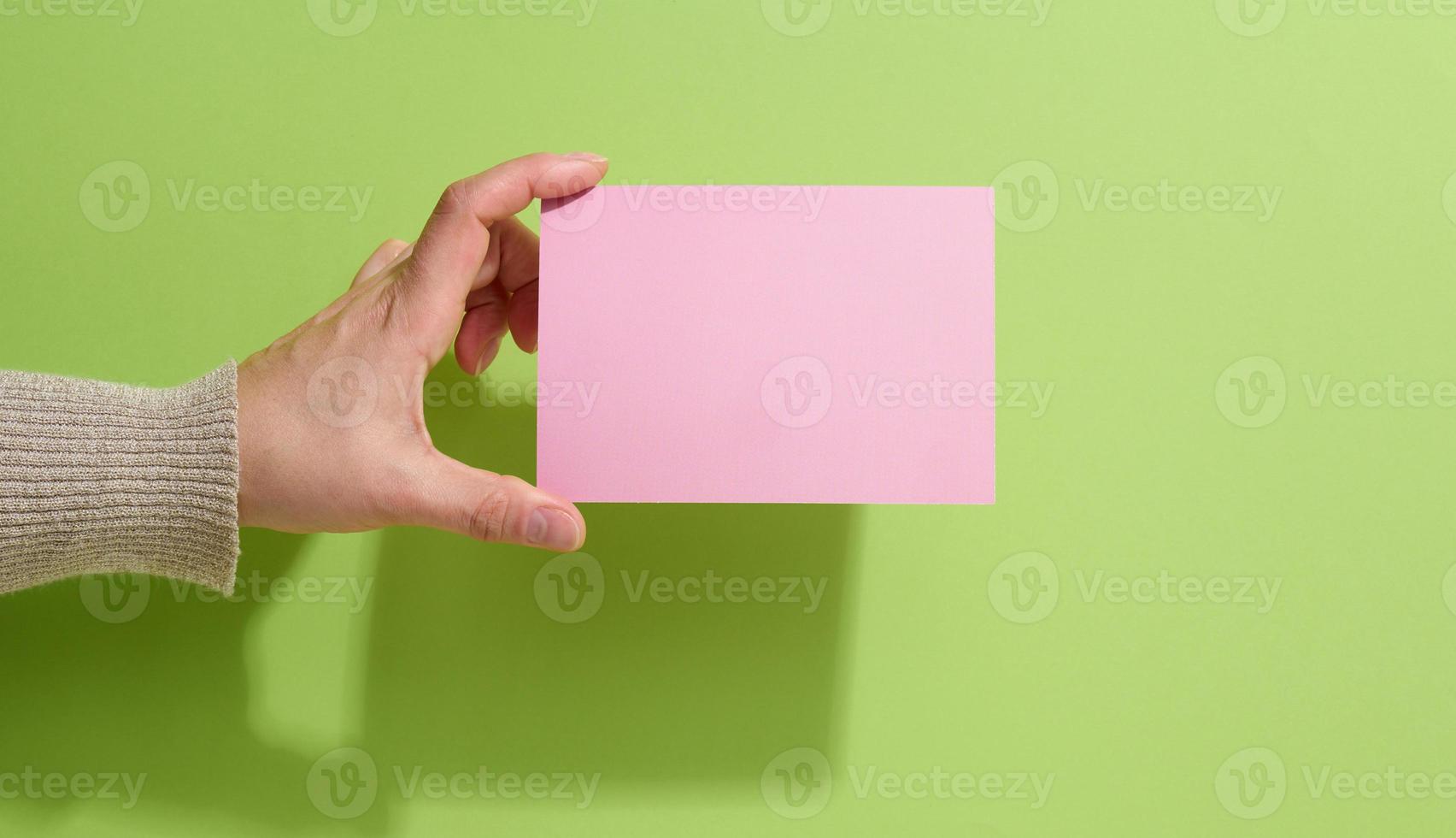 Female hand holding empty pink paper on a green background. Copy paste image or text, close up photo