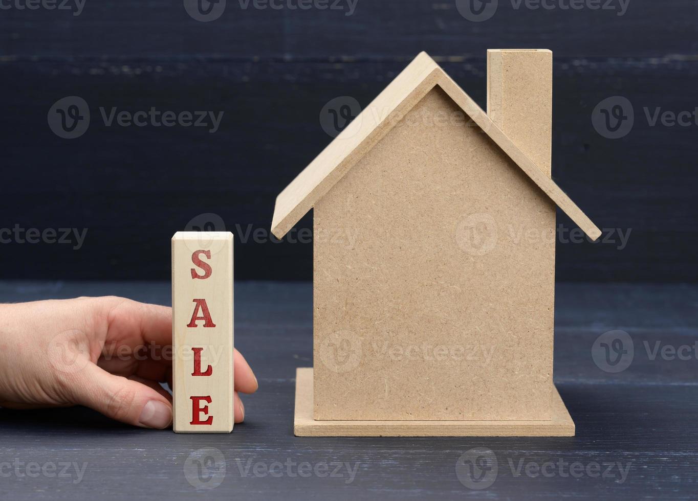 wooden model of a house and a hand holds a block with an inscription  sale on a blue background. House sale concept, real estate investment photo