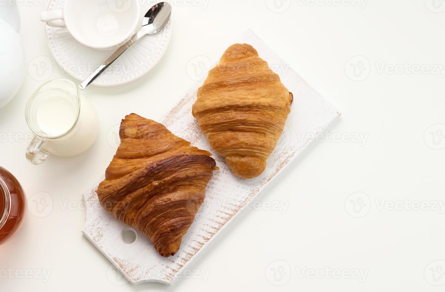 baked croissants, white ceramic teapot and empty cup and saucer, jar of honey on a white table, top view. Breakfast photo