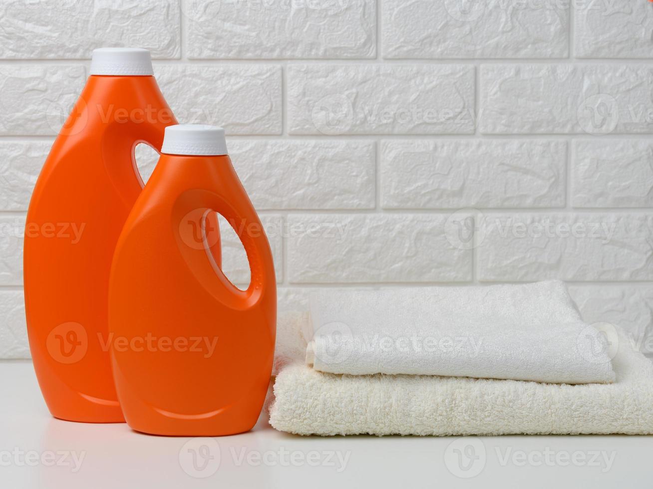 two orange plastic bottles of liquid detergent and a stack of towels on a white shelf, home wash photo