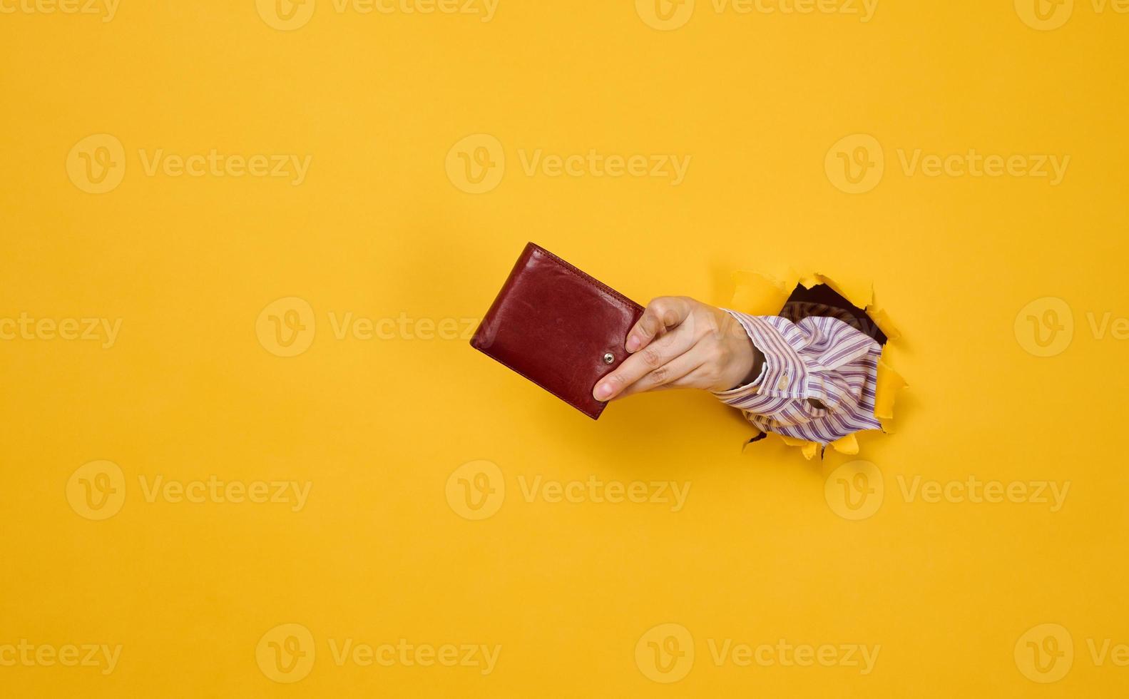 hand holds a brown leather wallet on a yellow background, part of the body sticks out of a torn hole in the paper photo