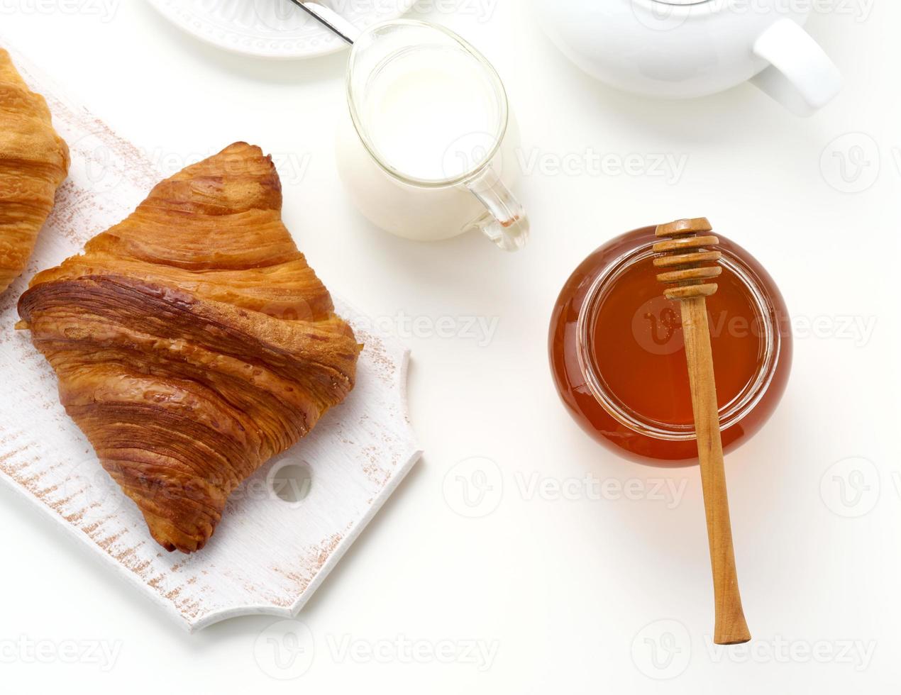 baked croissants, white ceramic teapot and empty cup and saucer, jar of honey on a white table, top view. Breakfast photo