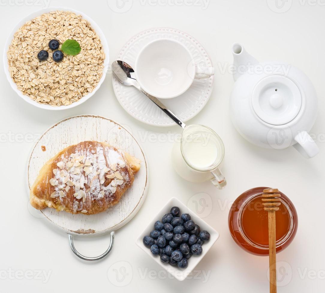 desayuno matutino, copos de avena crudos en un plato de cerámica, leche en un decantador, arándanos y miel en un frasco sobre una mesa blanca foto