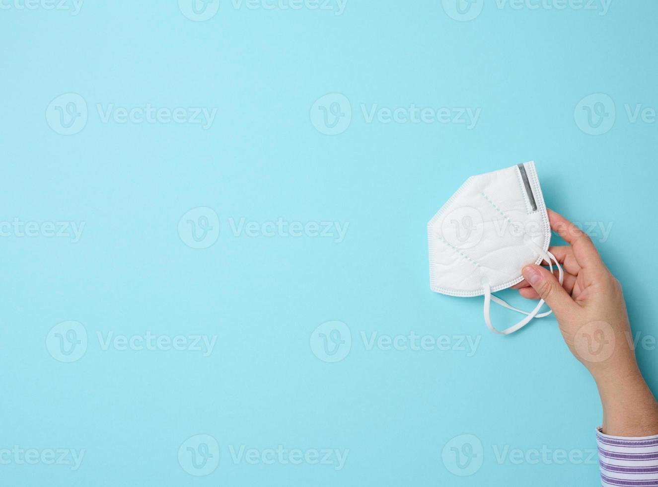 female hand holds white disposable medical face mask against viral diseases, blue background, top view photo